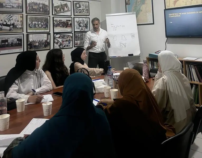 A diverse group of women participating in a community workshop at the Women-Adult-Youth (WAY) headquarters in Tripoli, Lebanon. A male facilitator stands by a whiteboard and screen titled 'Basic Elements of Art Composition' during an educational session