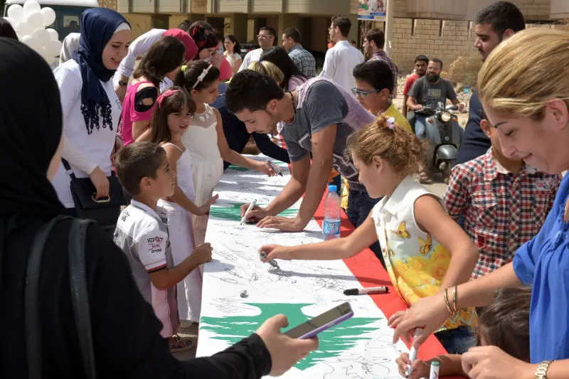 A vibrant outdoor community event by WAY Lebanon where children, youth, and adults gather around a long banner featuring the Lebanese cedar tree, actively drawing and writing messages together in Tripoli.